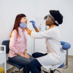 Young high skilled female afro ENT doctor checking woman's throat, larynx and oral cavity
