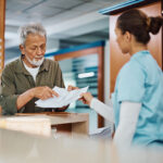 Senior man giving to nurse his medical paperwork while checking in at the clinic.