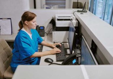 Caucasian middle aged woman working at medical reception desk typing on computer keyboard, sitting in modern healthcare facility, using office equipment and telephone