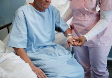 Elderly patient wearing hospital gown receiving comforting support from medical professional in pink scrubs holding patient's hand