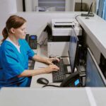 Caucasian middle aged woman working at medical reception desk typing on computer keyboard, sitting in modern healthcare facility, using office equipment and telephone