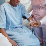 Elderly patient wearing hospital gown receiving comforting support from medical professional in pink scrubs holding patient's hand