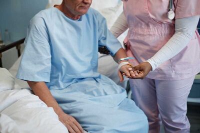 Elderly patient wearing hospital gown receiving comforting support from medical professional in pink scrubs holding patient's hand